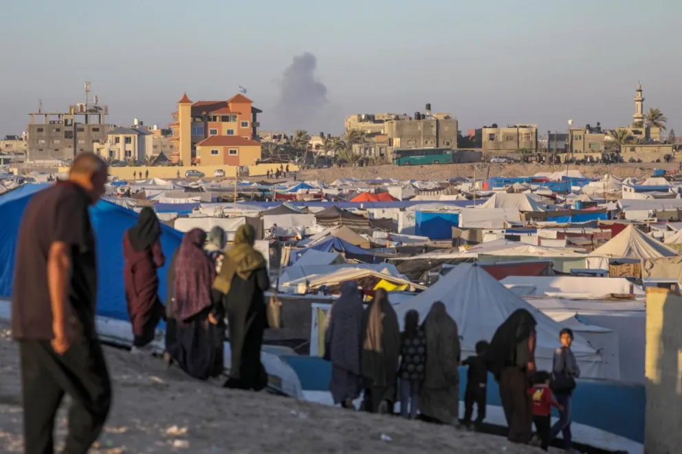 Desplazados palestinos en un campamento mientras se eleva humo al fondo tras un ataque aéreo israelí en Rafah. Foto: EFE