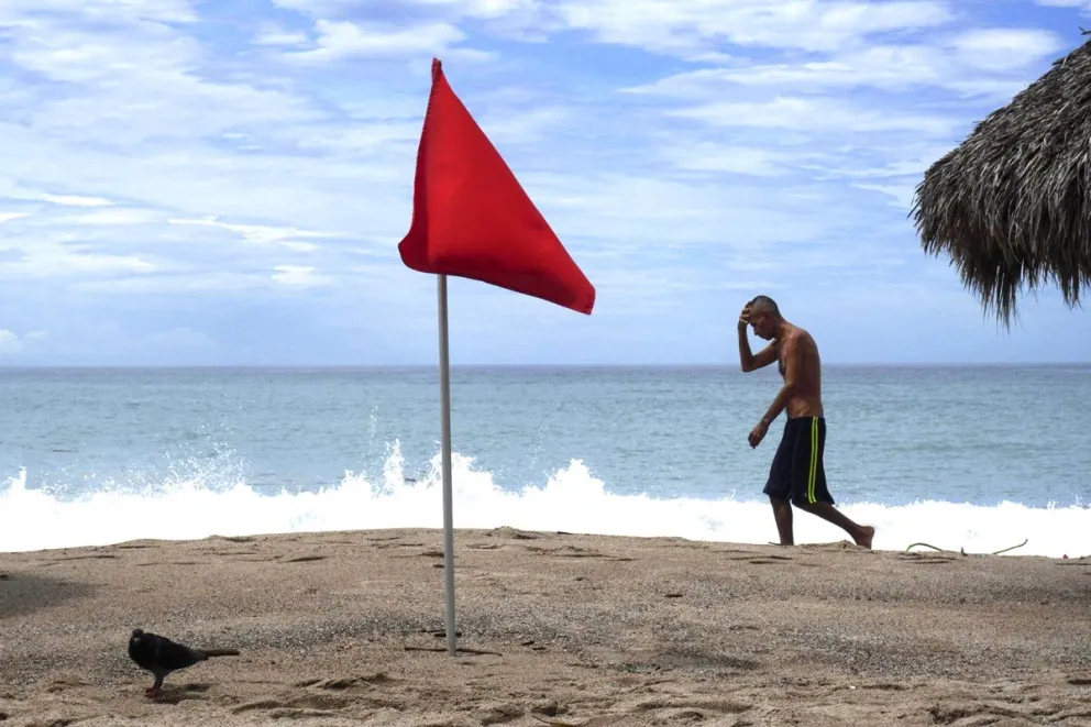 Un hombre camina cerca de una bandera roja como aviso del fuerte oleaje ante el paso del huracán Rachel. Foto: EFE